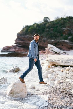 Kot pantolonlu ve kot ceketli bir adam yeşil kayalık dağların arka planında büyük bir taştan bir çakıl taşı kumsalına adım atıyor.