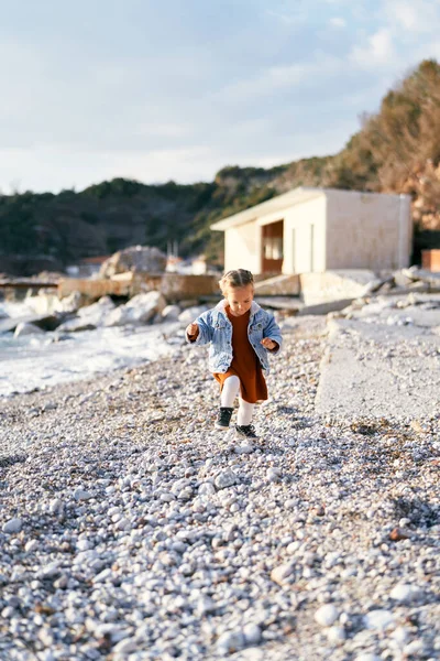 Маленькая девочка в платье и джинсовой куртке прогуливается по галечному пляжу, глядя на ноги на фоне моря, камней и здания — стоковое фото