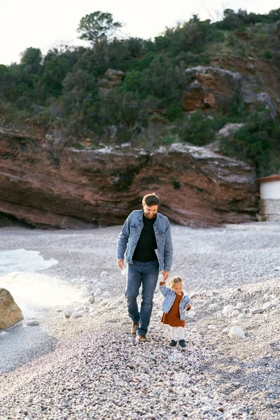 Улыбающийся папа ведет маленькую девочку за руку вдоль галечного пляжа на фоне зеленых скалистых гор — стоковое фото