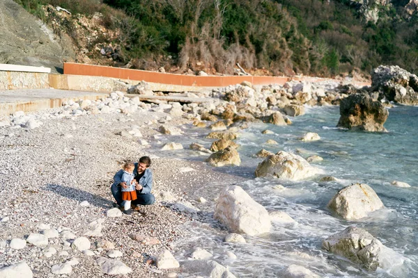 Улыбающийся папа сидел на корточках рядом с маленькой девочкой, раздавая камешки с каменистого пляжа у воды на фоне зеленых гор — стоковое фото