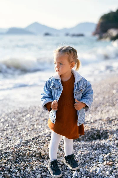 Gadis kecil yang termenung berdiri di pantai kerikil, berpegangan pada tepi jaket denim dan melihat ke kejauhan — Stok Foto