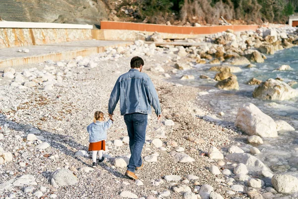 Папа в джинсовой куртке и джинсах держит маленькую девочку за руку, гуляя с ней вдоль скалистого берега у воды. Вид сзади — стоковое фото
