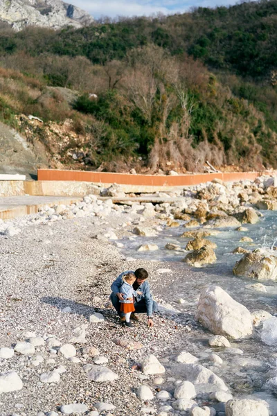 Папа сел на корточки рядом с маленькой девочкой, собирая камешки с каменистого пляжа у воды на фоне зеленой горы — стоковое фото