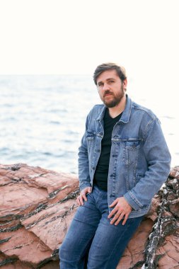 Kot pantolon ve kot ceketli dalgın bir adam denizin arka planında taşların üzerinde poz veriyor. Yan görünüm