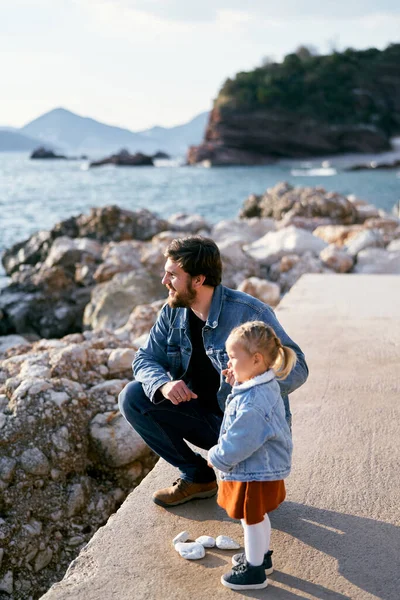 Smiling dad squatted next to a little girl on a large rock against the backdrop of rocky mountains. Dad and daughter are looking at the sea. Side view