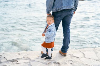 Kot ceketli ve kot pantolonlu bir baba, kayaların üzerinde suyun kenarında duran küçük bir kızın elini tutuyor. Kız yüzünü kameraya çevirdi.