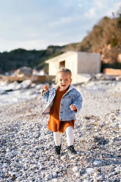 Gadis kecil tersenyum dengan gaun dan jaket denim berjalan di sepanjang pantai kerikil dengan latar belakang laut, batu dan bangunan — Stok Foto