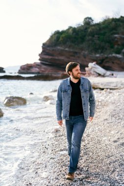 Kot pantolon ve kot ceketli gülümseyen adam çakıl taşı plajı boyunca yürür ve yeşil kayaların arka planında kıyıya bakar.