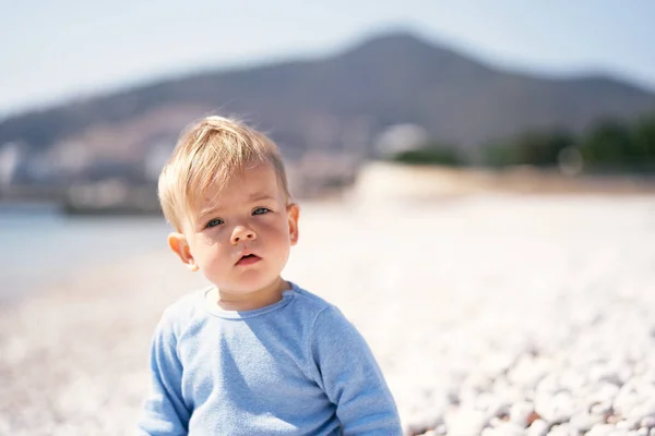 Komoly gyermek ül a kavicsos tengerparton a háttérben a hegyek. Közelkép. Portré — Stock Fotó