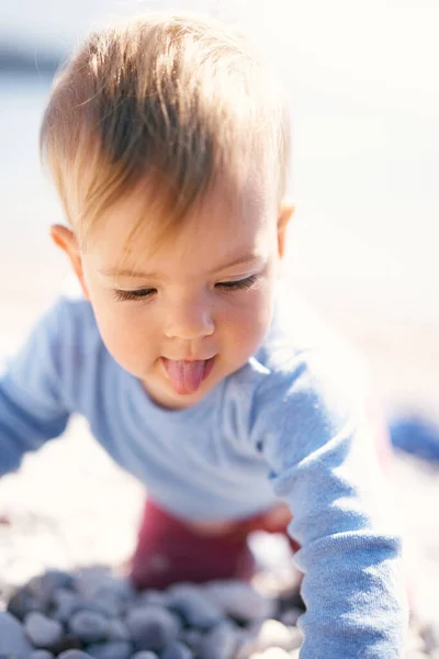 귀여운 아기가 혀를 내밀어 조약돌 해변에 무릎을 꿇고 앉아 있다. 클로즈업 — 스톡 사진