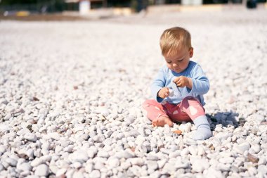 Mavi bluzlu şirin bir bebek ve kırmızı pantolon bir çakıl taşı kumsalında oturuyor, başını eğiyor ve ellerinde çakıl taşları tutuyor.