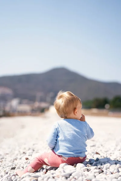 파란 스웨터를 입고 빨간 바지를 입은 아이는 작은 해변에 앉아 산을 배경으로 한다. 뒷 쪽을 봐. 클로즈업 — 스톡 사진