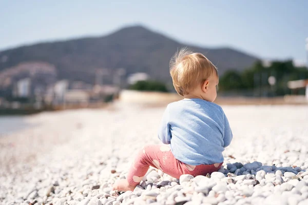 Anak dengan sweater biru dan celana merah duduk dengan kepalanya menoleh ke kanan di pantai kerikil dengan latar belakang pegunungan. Pandangan ke belakang. Close-up — Stok Foto