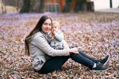 Gülümseyen anne parkta yaprakların üzerinde oturuyor kucağında tuttuğu sevimli bebeği kucaklıyor.