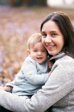 Gülümseyen anne, parkta yeşilliklerin üzerinde otururken gülümseyen bebeğe sarılıyor. Yakın plan. Portre