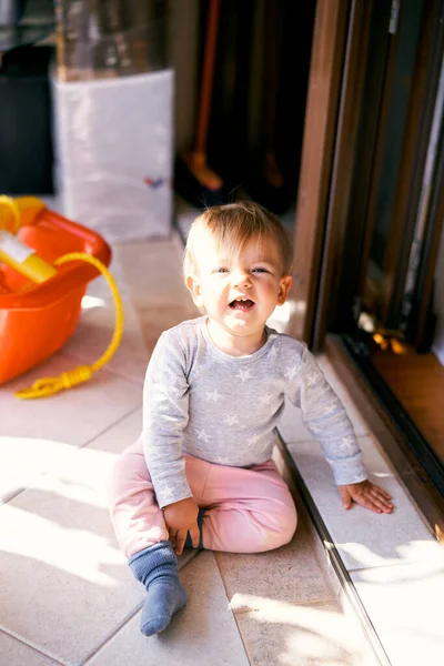 Smiling little child sitting on the floor on the balcony near the open door against the background of an orange bowl with a clothesline — Stock Photo, Image