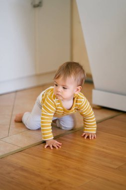 Çizgili bluz giymiş bir bebek ve beyaz mobilyaların arka planında fayanslardan plastik kaplamalara kadar sürünüyor.