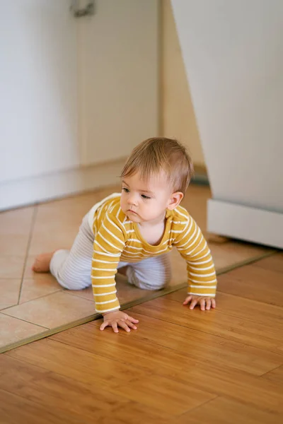 Baby i randig blus och byxor kryper från en kakel till ett laminat i köket mot en bakgrund av vita möbler — Stockfoto