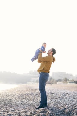 Kahverengi ceketli ve kotlu gülümseyen baba, kumsalda dururken mavi tulumlu küçük bir çocuğu kollarında tutuyor.