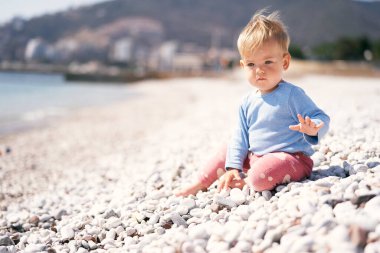 Ciddi çocuk, elini kaldırmış deniz kenarındaki bir çakıl taşlı plajda, dağların arka planında oturuyor.