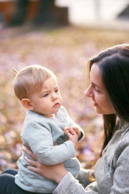 Gülümseyen anne bebeği kucağında tutuyor, yüz yüze, parkta yaprakların üzerinde oturuyor. Yakın plan.