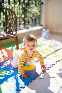 Çocuk, balkondaki renkli bir halıda hasır bir sandalyenin arka planında sürünüyor.
