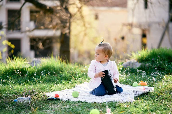 Liten flicka sitter på en filt bland leksaker på grönt gräs och håller en leksaksbil i händerna — Stockfoto