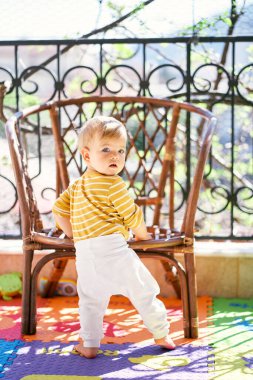 Çocuk hasır sandalyenin yanındaki balkonda duruyor, başını çeviriyor.