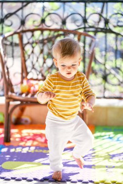 Küçük bir çocuk balkondaki hasır bir sandalyenin arka planında renkli bir halıda yürüyor.