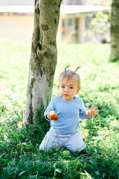 Petite fille s'assoit à genoux sous un arbre dans l'herbe verte et tient une pêche dans sa main — Photo