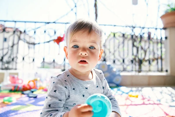 Bayi lucu duduk di karpet berwarna di balkon dan memegang mainan plastik di tangannya — Stok Foto
