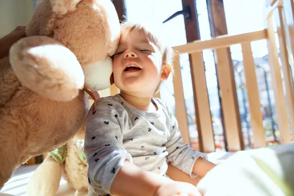 Tersenyum anak kecil duduk di tempat tidur kayu bersandar terhadap boneka beruang besar — Stok Foto