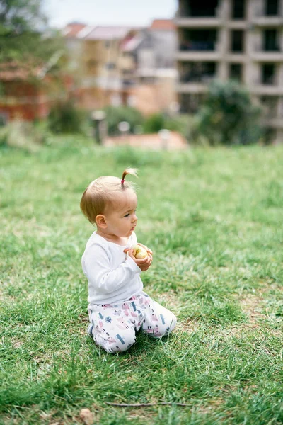 Niña sentada en un prado verde con una manzana en la mano sobre el fondo de una casa —  Fotos de Stock