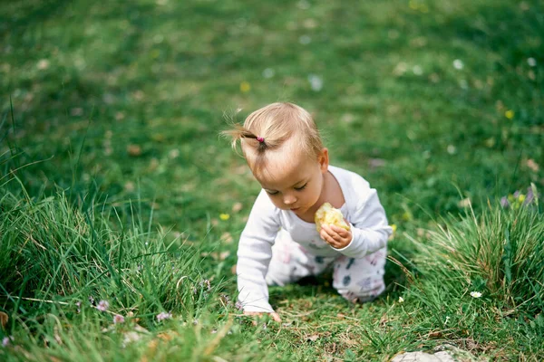 Liten flicka sitter på knä lutande sig fram på en grön äng med ett äpple i handen — Stockfoto