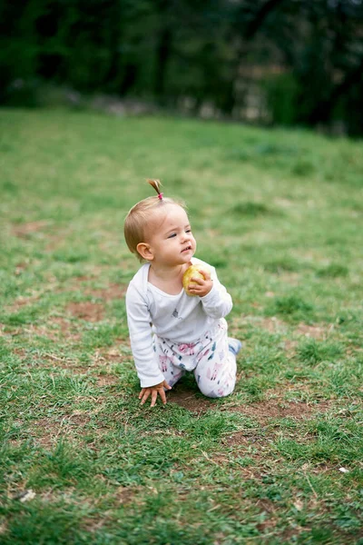 La bambina si inginocchia su un prato verde con una mela in mano — Foto Stock