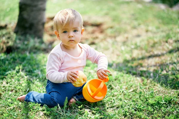 Little girl sits on a green lawn and holds a yellow bucket and an apple in her hands — Stock Photo, Image