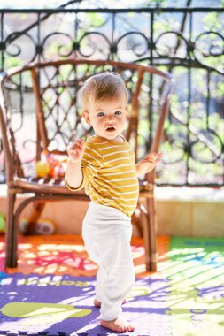 Küçük çocuk balkonda hasır bir sandalyenin önünde duruyor.