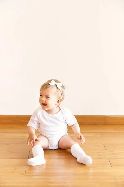 Liten skrattande flicka med en båge på huvudet sitter på golvet i rummet — Stockfoto