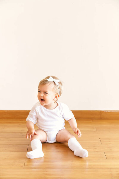 Little laughing girl with a bow on her head sits on the floor in the room