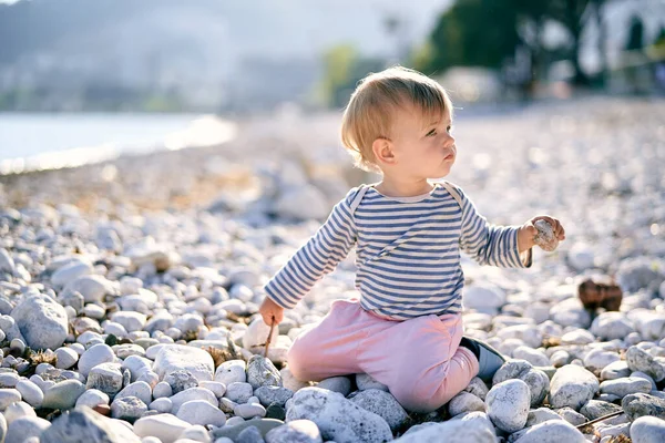 아이는 무릎을 꿇고 앉아서 자갈 해변에서 머리를 돌려 손에 조약돌을 쥐고 있습니다 — 스톡 사진