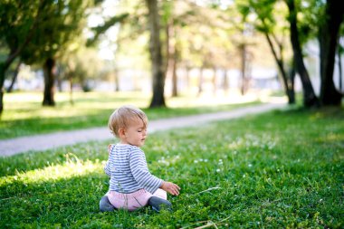 Küçük bir çocuk, yeşil bir çimenlikte, kaldırımın kenarında başı dönük oturuyor. Arka plan