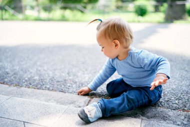 Parktaki yolda oturan ve parmağıyla fayansları kaşıyan küçük kız.