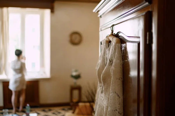 A bride in a robe stands at the window in a hotel room, a wedding dress hangs on the wardrobe door — Stock fotografie