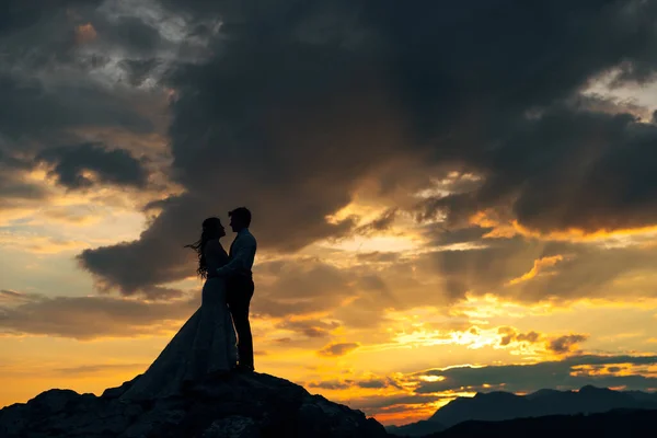 The bride and groom are embracing on the rocks in the mountains at sunset, silhouettes — Φωτογραφία Αρχείου