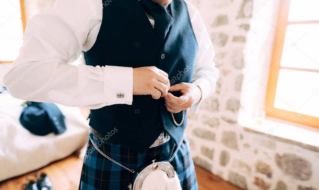 A man in Scottish national dress buttons up his waistcoat and prepares for a wedding ceremony in a hotel room, close-up 