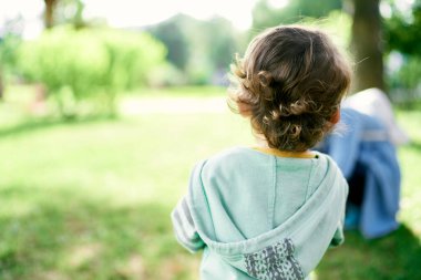 Yeşil çimlerin üzerinde kapüşonlu kazaklı kıvırcık saçlı çocuk duruyor. Arka plan