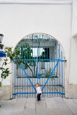 Çocuk evin avlusunda duruyor ve elini kafes kapısına dayıyor.