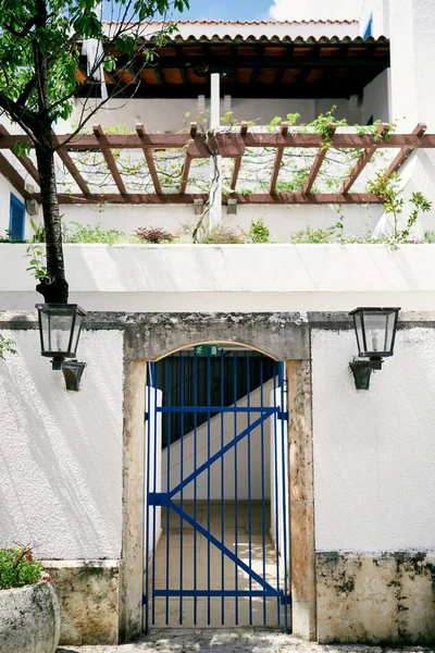 Sten hegn med gitter porte nær huset med en balkon, træ og grønt - Stock-foto