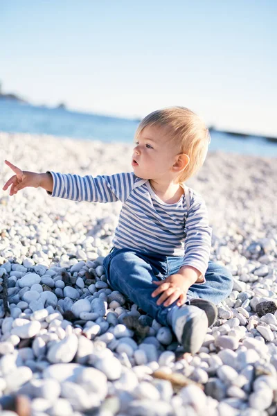 Anak itu duduk di pantai kerikil di tepi laut dan menunjuk jarinya ke kejauhan — Stok Foto