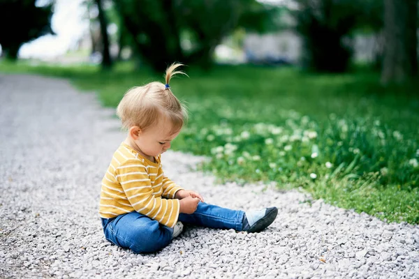 Kleines Mädchen mit Pferdeschwanz auf dem Kopf sitzt auf einem Kiesweg in einem grünen Park — Stockfoto